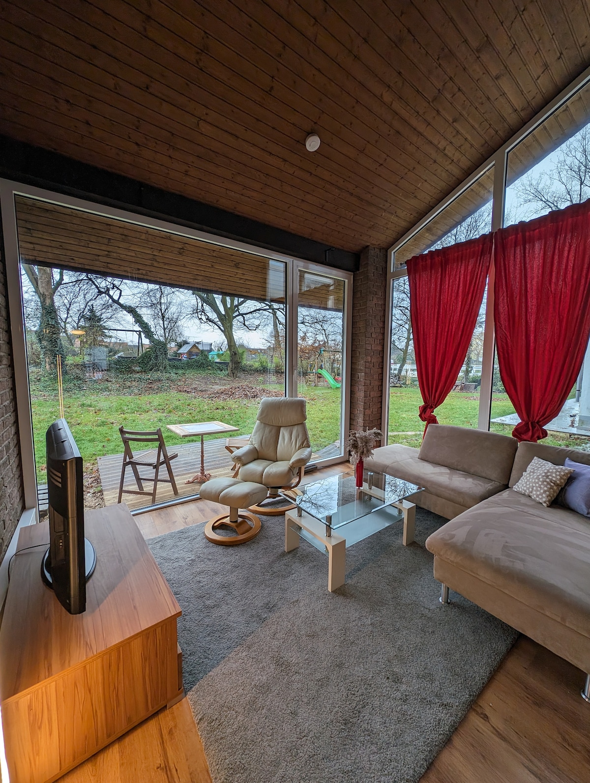 A modern living area showcases large glass windows that invite natural light and offer a view of the lush garden. A soft couch is paired with a stylish armchair and a small coffee table, while red curtains add a pop of color to the neutral tones.