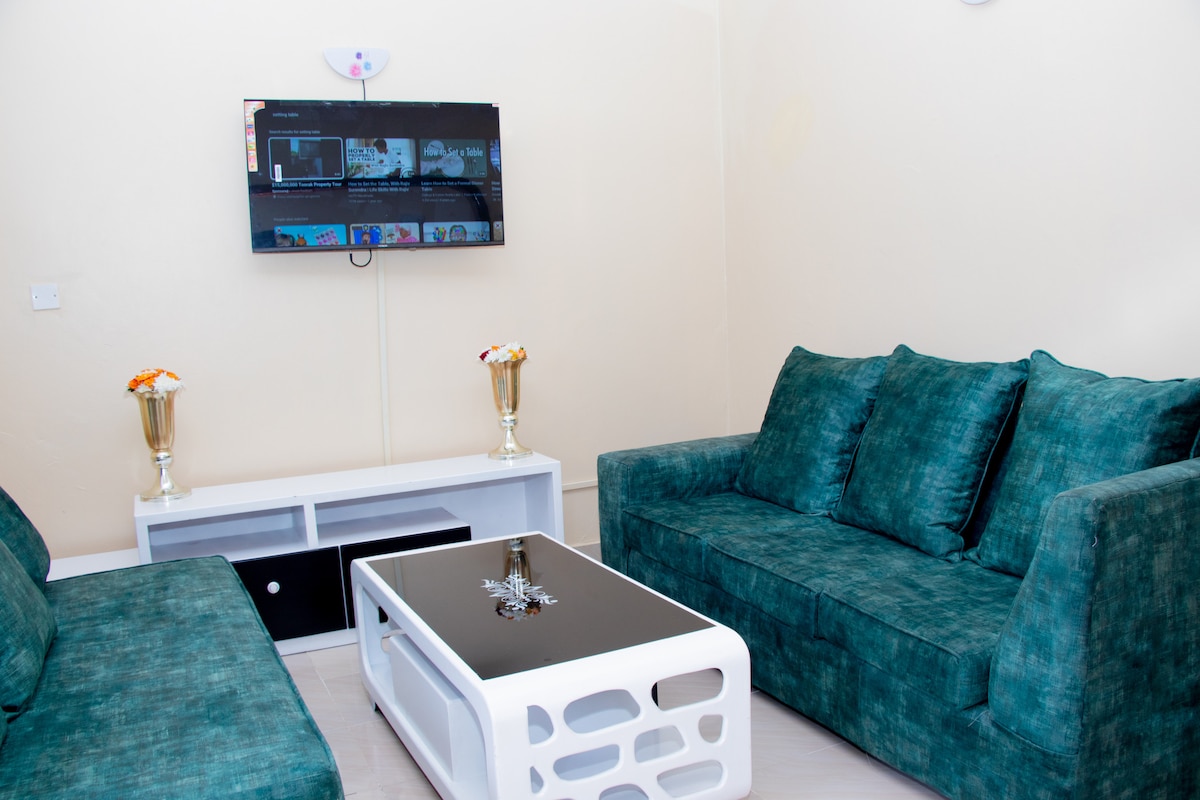 A comfortable living area is presented, featuring a modern black coffee table surrounded by plush teal sofas. A wall-mounted television is visible, offering entertainment options. Decorative vases with flowers add a subtle touch to the space, enhancing its inviting character.