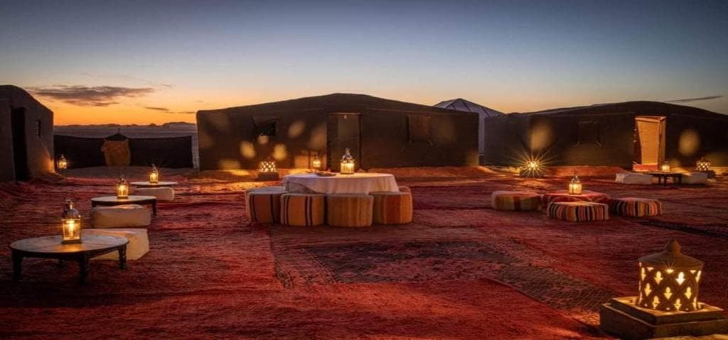 An outdoor seating area is illuminated by soft lantern light, casting gentle reflections on the surrounding desert landscape. Spacious seating arrangements, including low tables and cushions, are arranged on rich red carpeting, creating a welcoming space for relaxation under the evening sky.
