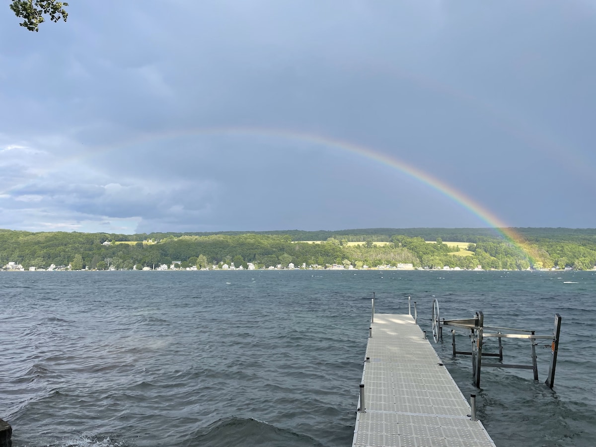 The image captures a wooden dock extending towards Keuka Lake, with gentle waves lapping at its edge. A prominent rainbow arches across the sky, contrasting with the gray clouds. Lush green hills frame the background, suggesting a serene lakeside environment.