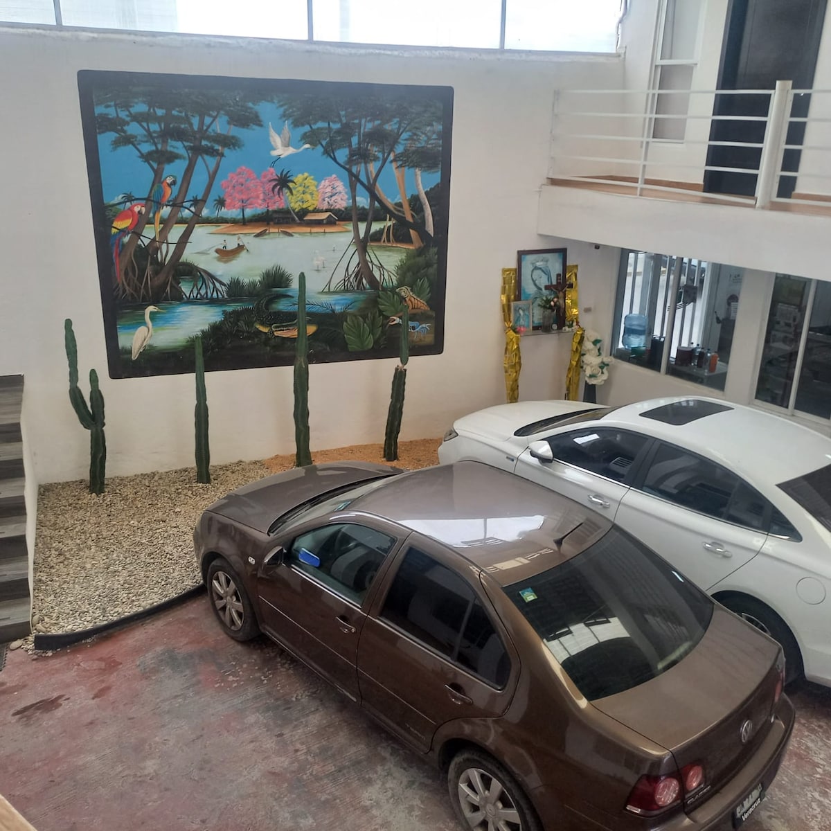 A spacious parking area is shown, featuring a large mural depicting a serene outdoor scene with tropical plants and birds. Two cars are parked on a surface of red and gray flooring surrounded by decorative stones and cacti adding a natural element.