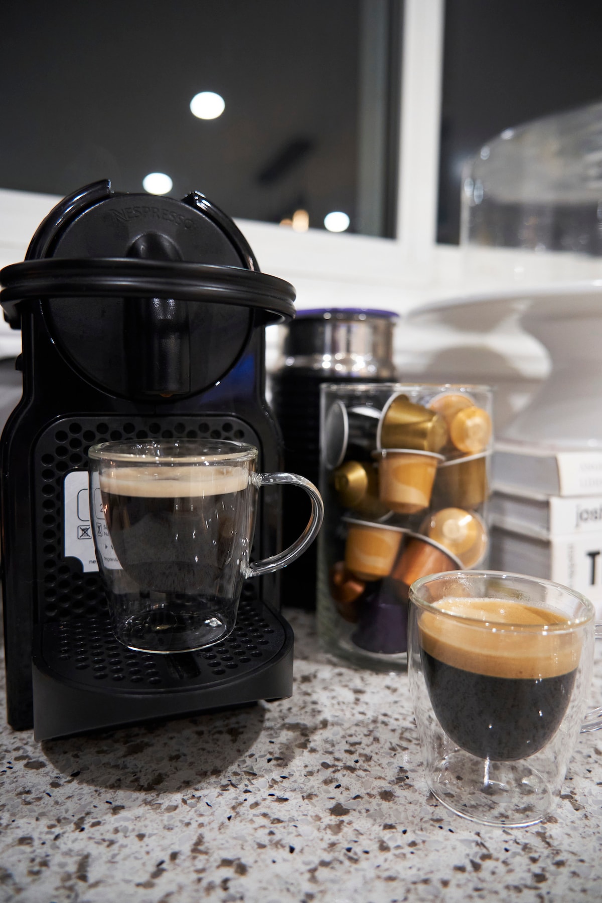 A modern coffee machine sits on a speckled countertop, with two cups of coffee positioned in front. A clear container with colorful coffee capsules is visible beside it, adding a touch of organization to the setting.