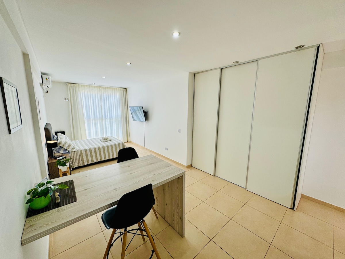 A bright apartment interior is seen, featuring a spacious bed with striped bedding across from a desk and a chair. A large window allows natural light to flood the room, while a minimalist closet occupies one wall. A television is mounted on the opposite wall.