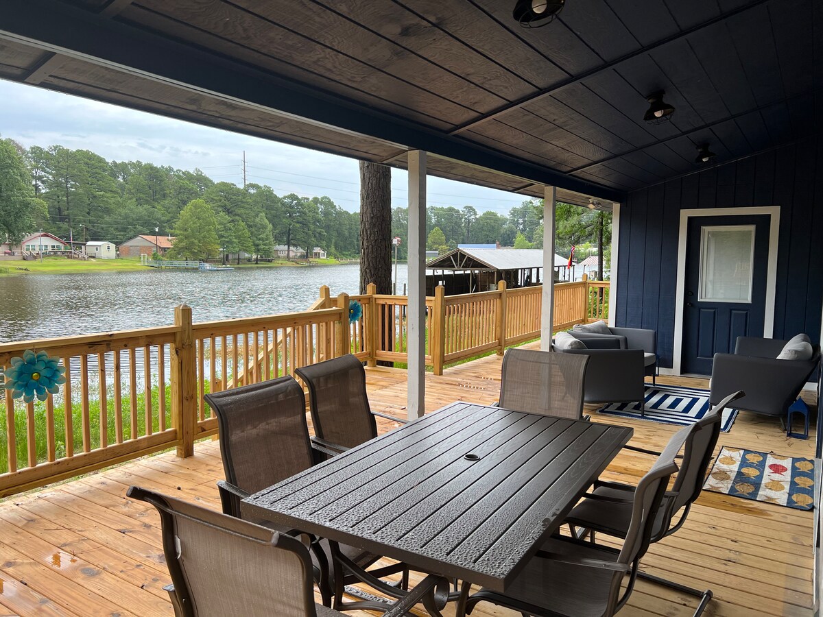 An outdoor deck is seen overlooking a serene lake, surrounded by green trees. A table with multiple chairs is positioned near the railing, providing a space for dining. Comfortable seating areas with soft cushions are arranged for relaxation, enhancing the lakeside experience.