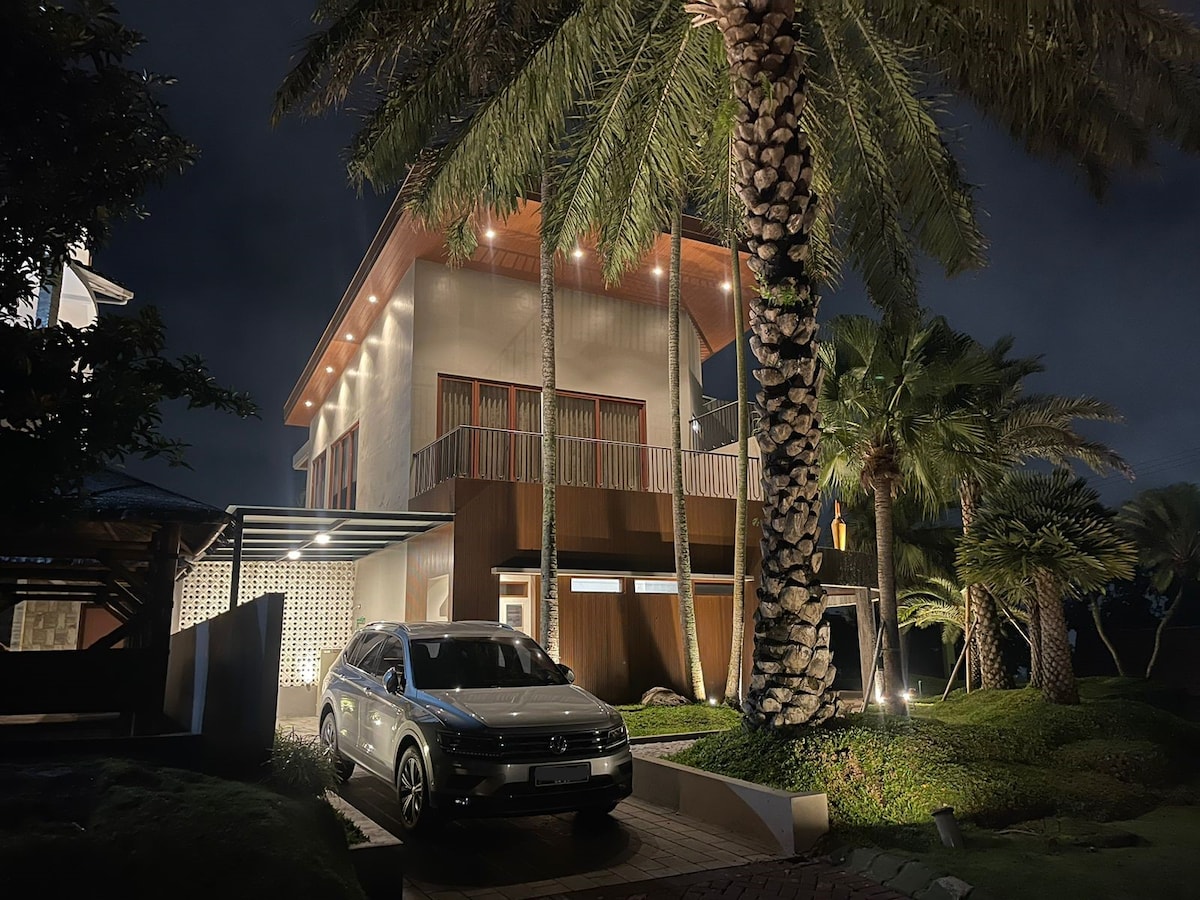 The villa exterior is showcased at night, featuring a modern structure surrounded by palm trees. A parked car is positioned in the driveway, illuminated by soft outdoor lighting, highlighting the contemporary architecture and lush landscaping.