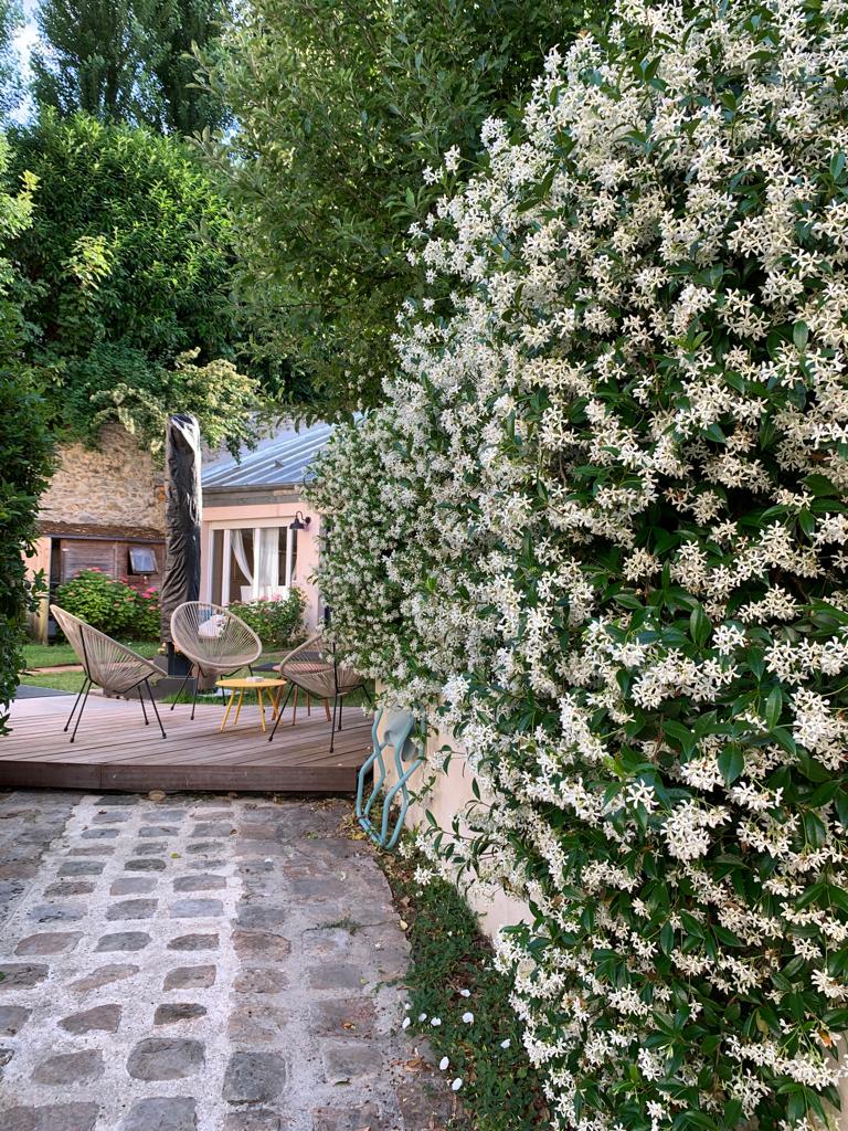 A charming outdoor area is surrounded by lush greenery and blooming white flowers. A wooden deck features comfortable seating arrangements, including woven chairs, creating an inviting space for relaxation. The scenic garden view enhances the tranquil atmosphere enjoyed from this cozy nook.