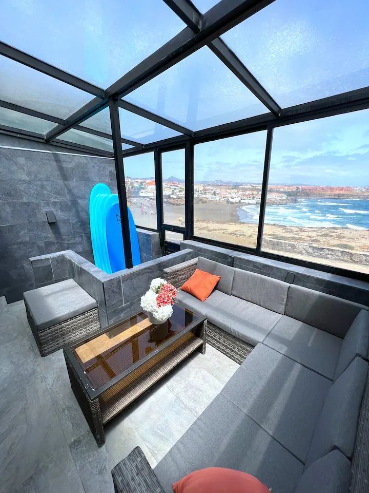A sunlit lounge area is enclosed by glass walls, offering panoramic views of the nearby beach. Gray upholstered seating is arranged around a coffee table, while decorative cushions add a touch of color. Surfboards are visible, suggesting opportunities for water activities.
