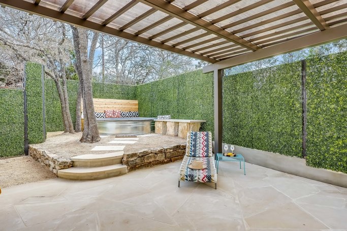 A gated side patio features a spacious stone terraced area with a covered seating option. Lush greenery lines the walls, and a Cowboy Pool is visible in the corner, accompanied by an outdoor dining area and a BBQ grill. Sunlight filters through the pergola.