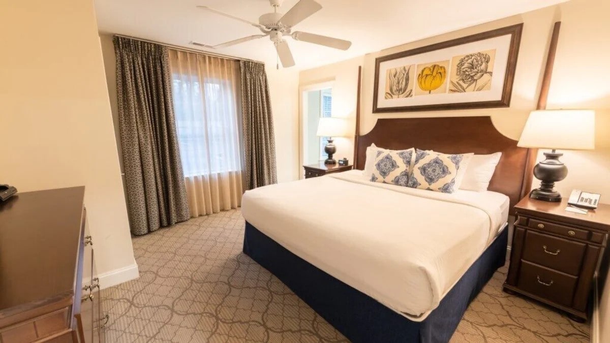 A well-appointed bedroom is presented with a spacious bed, complemented by bedside tables featuring lamps. Large windows are draped with sheer and patterned curtains, allowing soft light to filter in. Artwork highlighting floral designs decorates the wall above the bed.