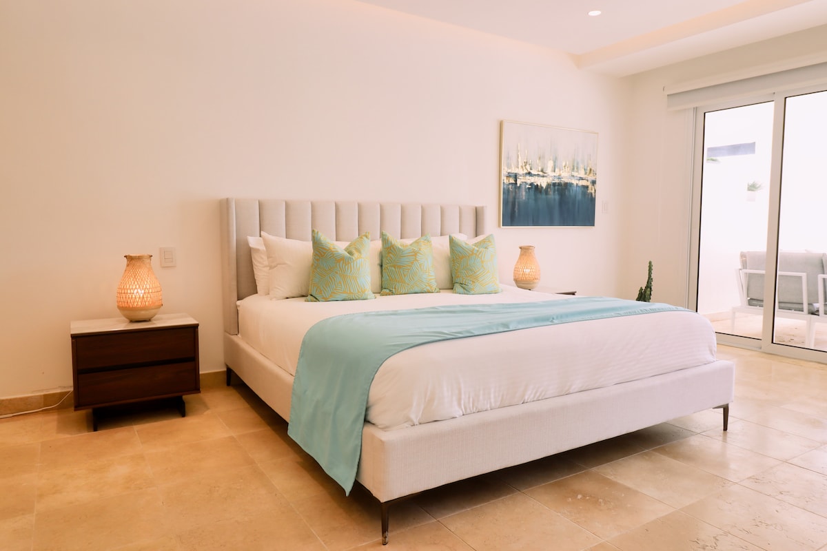A spacious bedroom features a large bed with decorative pillows and a light blue throw. Two bedside lamps sit on wooden tables, providing gentle illumination. Natural light filters in through glass doors that open to a cozy outdoor area, enhancing the room's bright and airy feel.