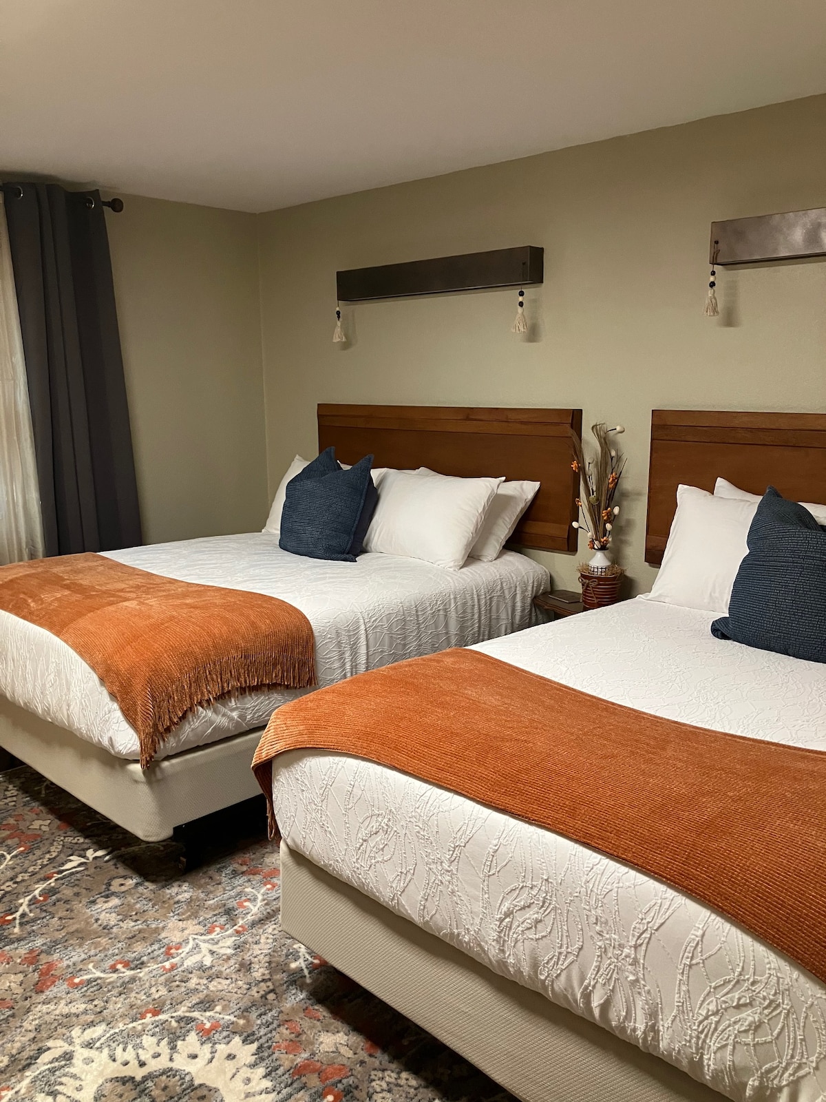 A guest room features two spacious beds with light linens and decorative throw blankets in warm tones. Each bed is framed by wooden headboards. A decorative arrangement is placed between the beds, and large windows are covered with dark curtains, allowing for natural light.
