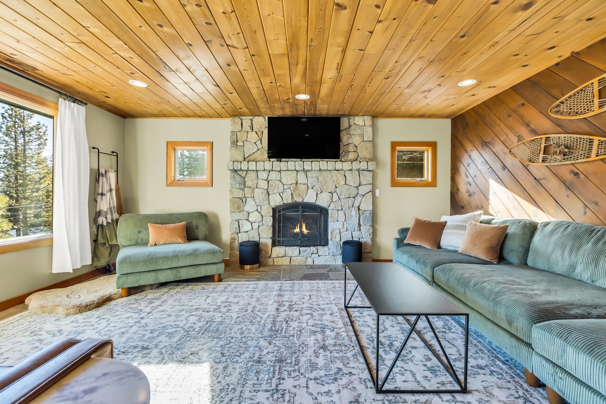 A cozy living space features a large sectional couch with cushions, a loveseat, and a rug covering the wooden floor. A stone fireplace is centered against a wooden accent wall, with a TV mounted above. Natural light enters through windows with views of the surrounding trees.