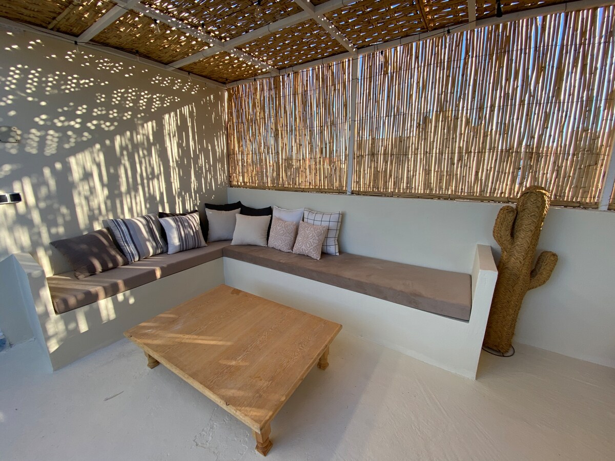 A cozy lounge area is highlighted by a built-in, cushioned seating space adorned with various throw pillows. A wooden coffee table is centered on the light-colored floor. Natural light filters through the bamboo ceiling and woven wall, creating a warm, inviting atmosphere.