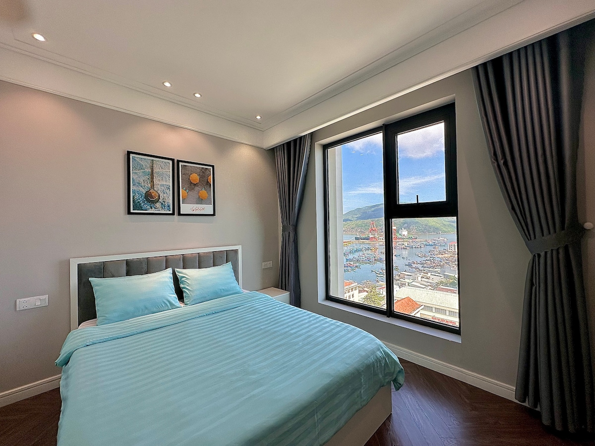 A well-lit bedroom features a spacious bed with soft blue bedding, complemented by two framed artworks on the walls. Large windows offer a view of the harbor and surrounding landscape, with gray curtains elegantly framing the window.