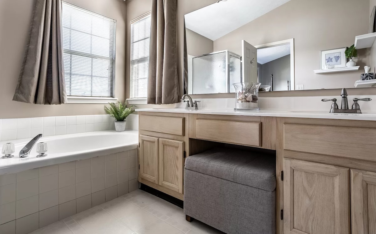 A well-lit bathroom features a spacious vanity with natural wood cabinetry and a sink. A soaking tub is placed next to a window with soft curtains. A shower area is visible in the background, with a light-colored tile floor enhancing the inviting space.