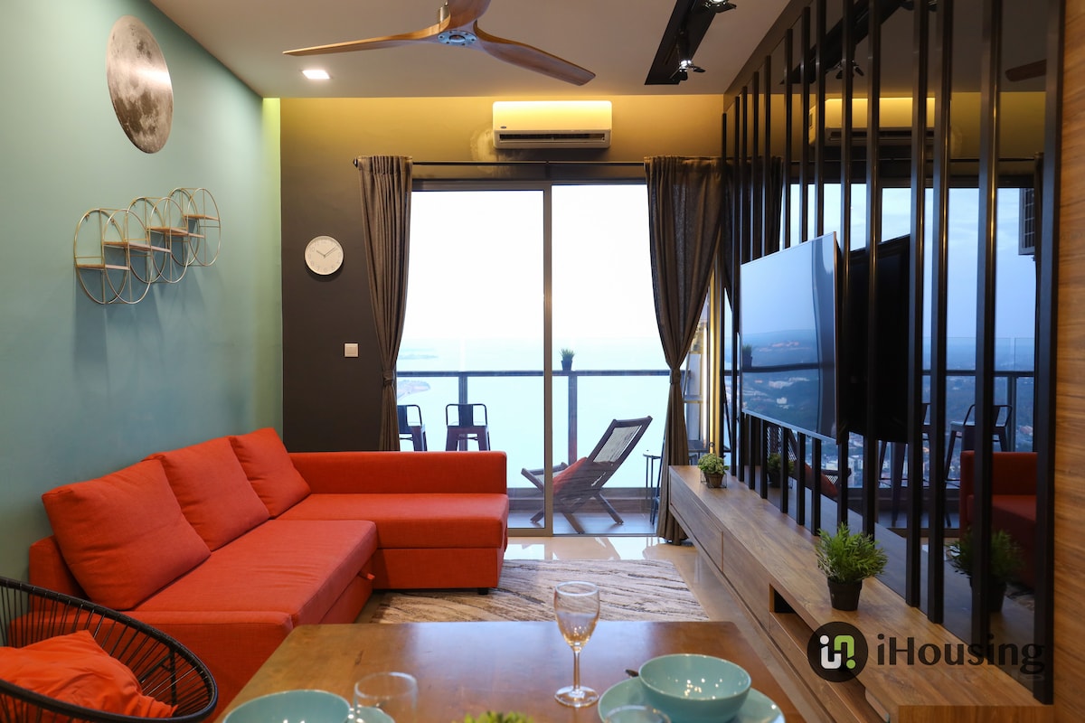 A modern living room features a red sofa facing a wall-mounted television. Floor-to-ceiling windows offer a view of the sea, with sliding glass doors leading to a balcony. A round dining table with place settings and decorative items completes the inviting space.