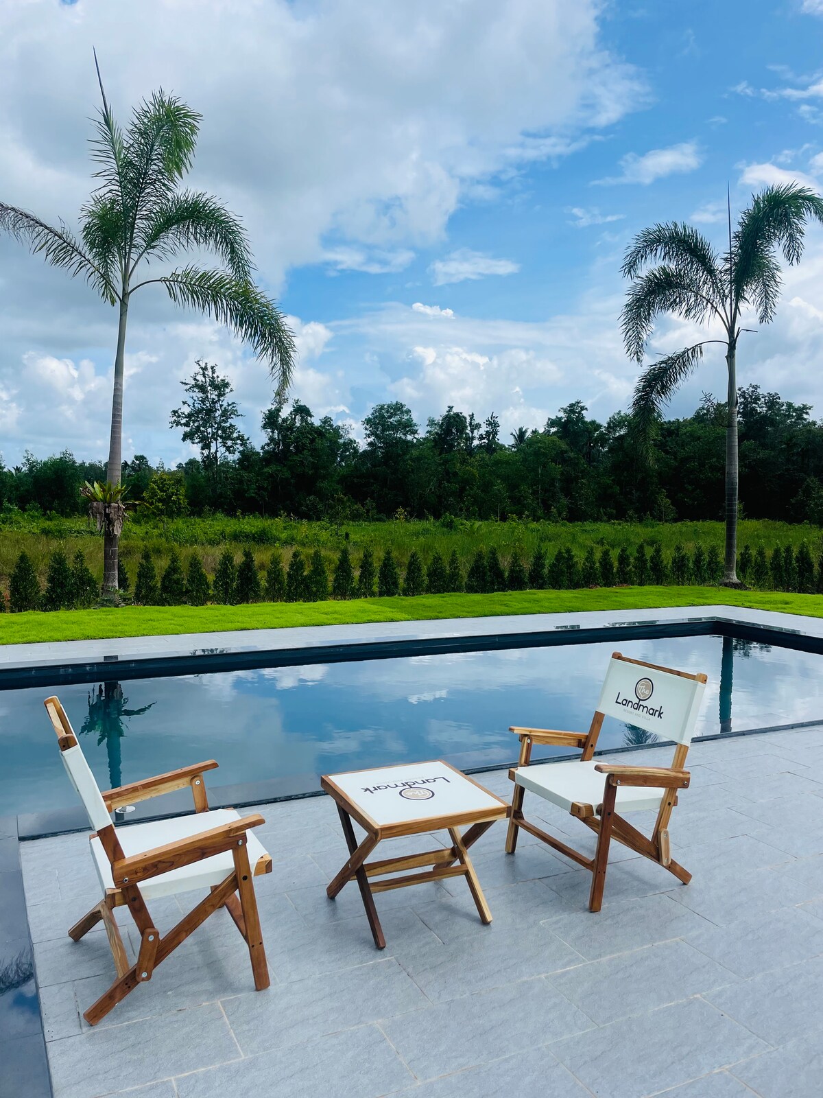 Two wooden chairs with white fabric are positioned next to a serene pool, reflecting the clear sky. A small table sits between them on a grey stone patio, surrounded by lush greenery and palm trees, creating a calm outdoor setting.