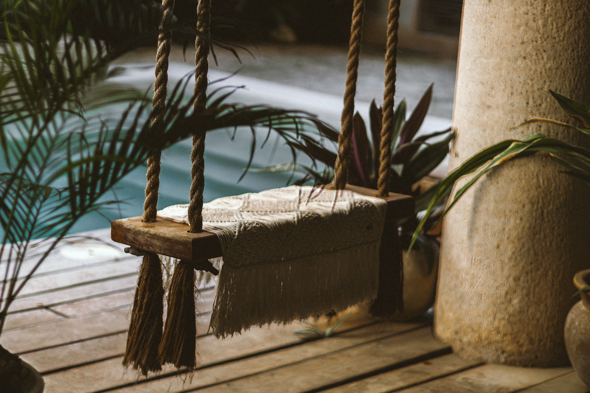 A rustic wooden swing is suspended from thick ropes, surrounded by lush green plants. Textured fabric drapes over the swing, offering a cozy touch. In the background, a serene pool is visible, contributing to the tranquil atmosphere of the space.