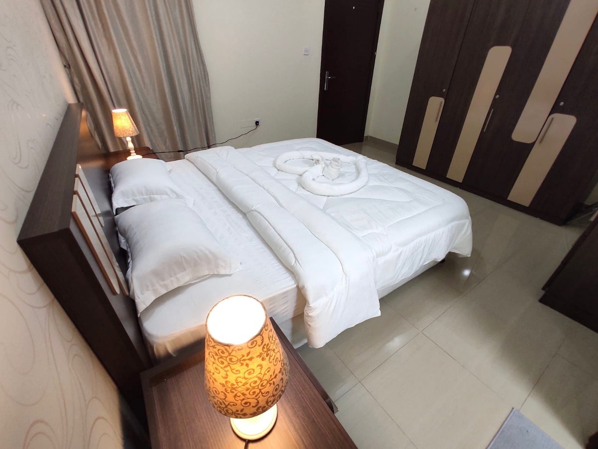 A well-arranged bedroom features a spacious bed with fresh linens and decorative pillows. A lamp casts a warm glow beside the bed, while a wardrobe with striped accents is visible in the background. Soft curtains gently frame a window, enhancing the overall comfort.