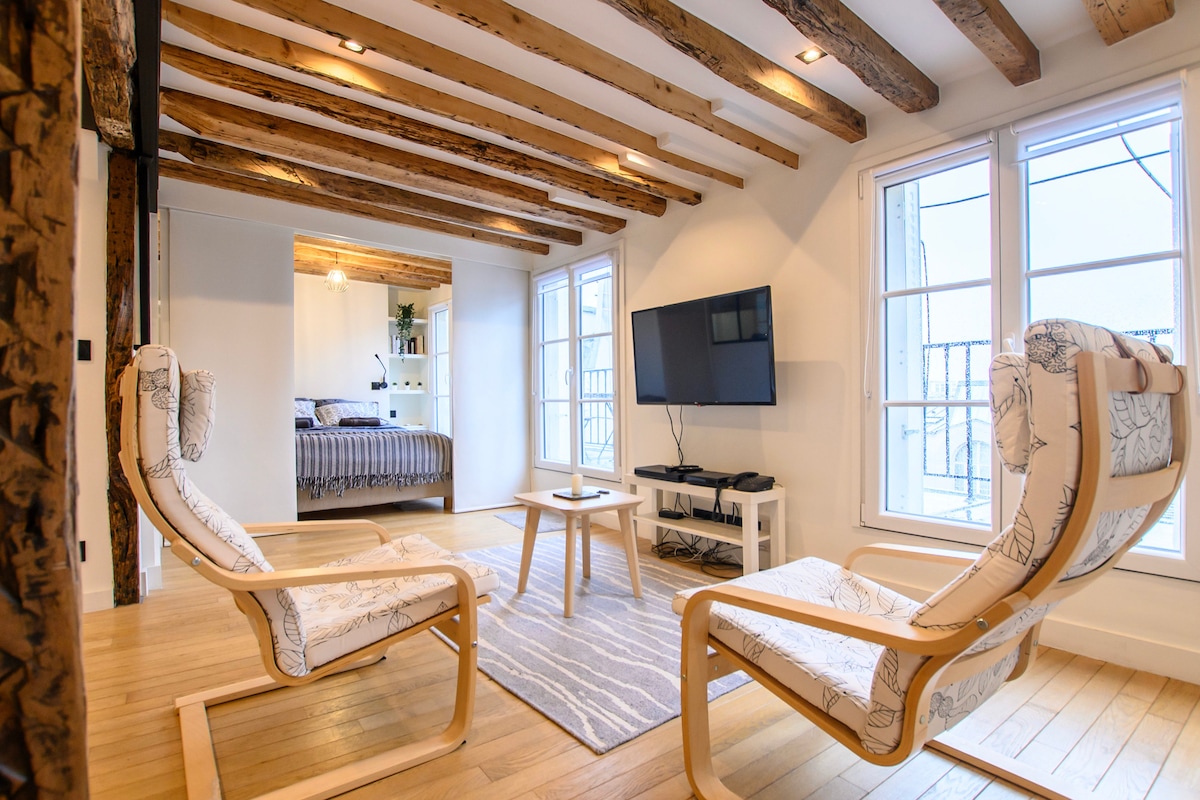 A bright living area features two modern armchairs positioned around a small wooden table, with a neutral area rug underfoot. Large windows let in natural light, showcasing the wooden beams of the ceiling and providing a view into a connected sleeping area.