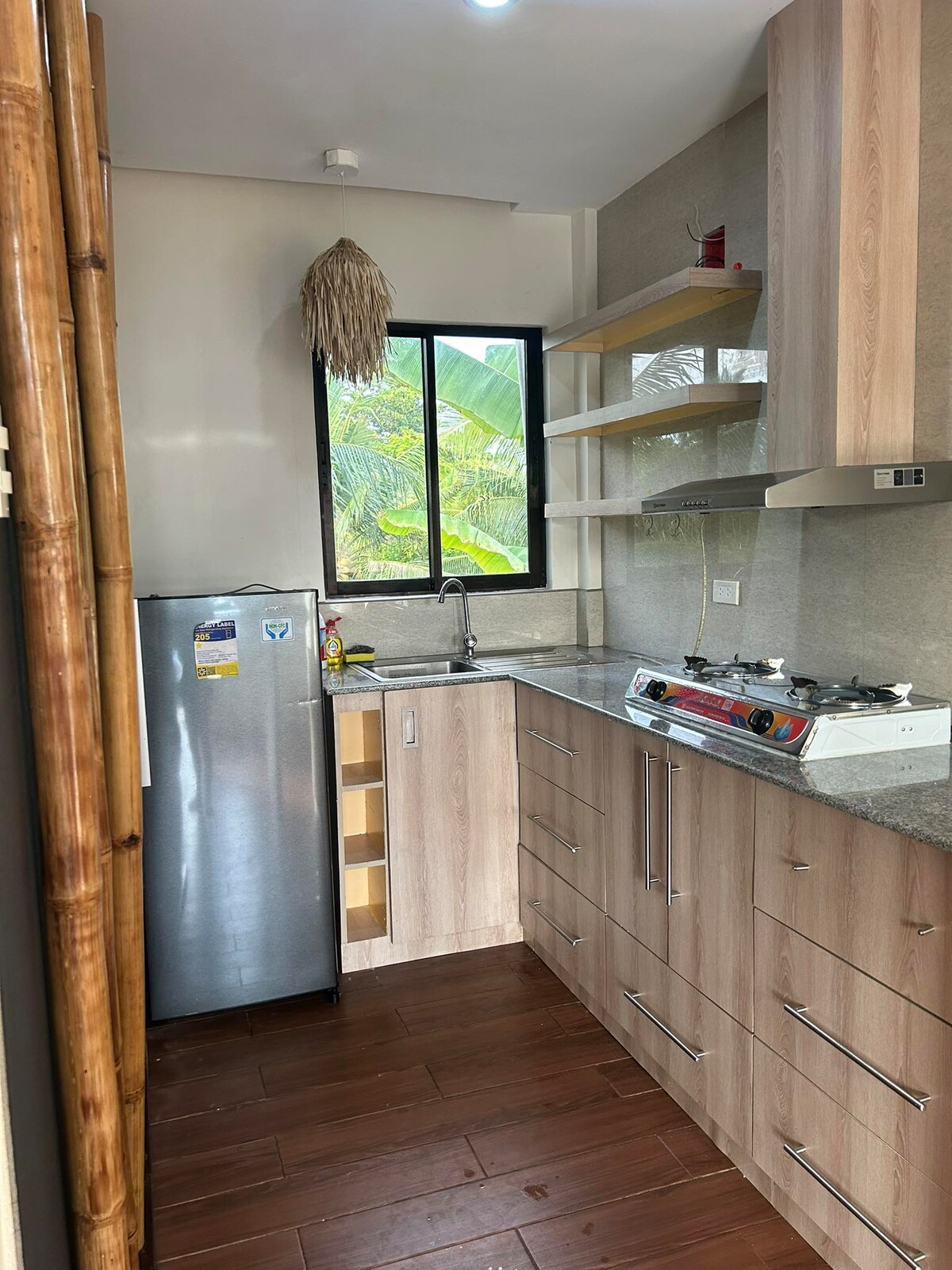 A modern kitchen space is presented, featuring wooden cabinetry, a stainless steel refrigerator, and a gas stovetop. Natural light streams in through a large window, showcasing lush greenery outside. Counter surfaces are finished in a light stone texture, enhancing the contemporary design.