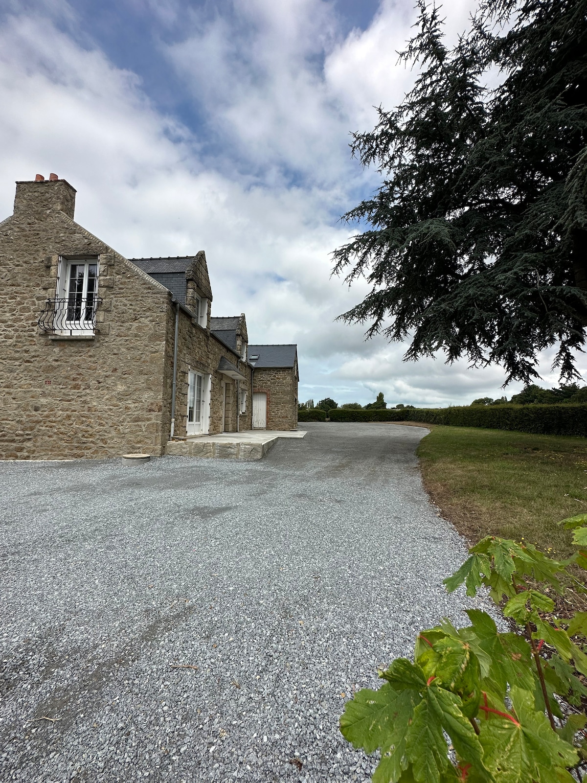 A gravel pathway leads to a rustic stone building, showcasing a spacious exterior surrounded by greenery. A tree provides shade in the foreground. The sky is partly cloudy, creating a serene atmosphere in the countryside setting.
