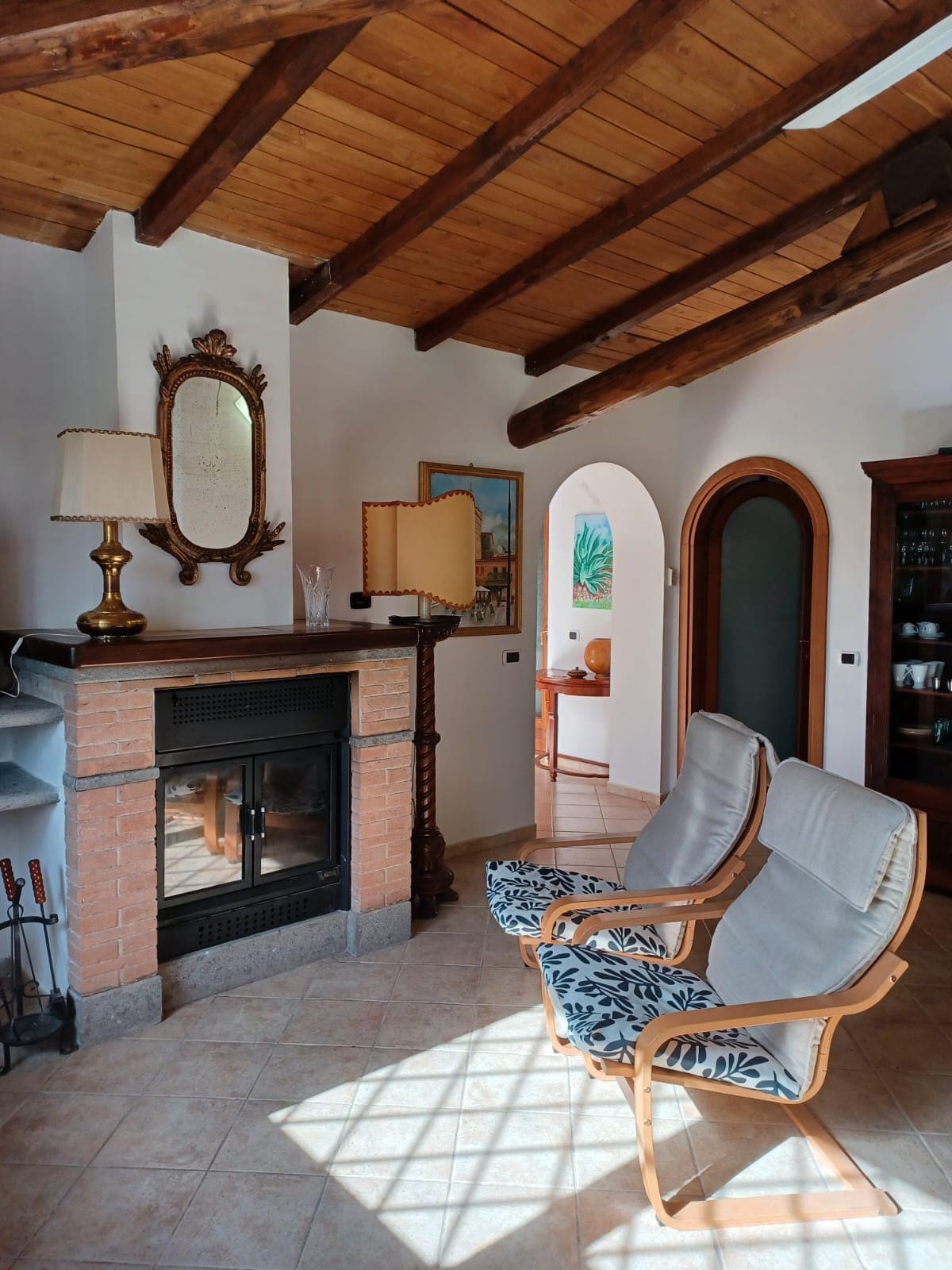 A cozy living area features a brick fireplace, accented by a wooden beam ceiling. Two light-colored armchairs are positioned nearby, creating a relaxed seating arrangement. Natural light filters through a nearby arch, highlighting the neutral tones and inviting decor.