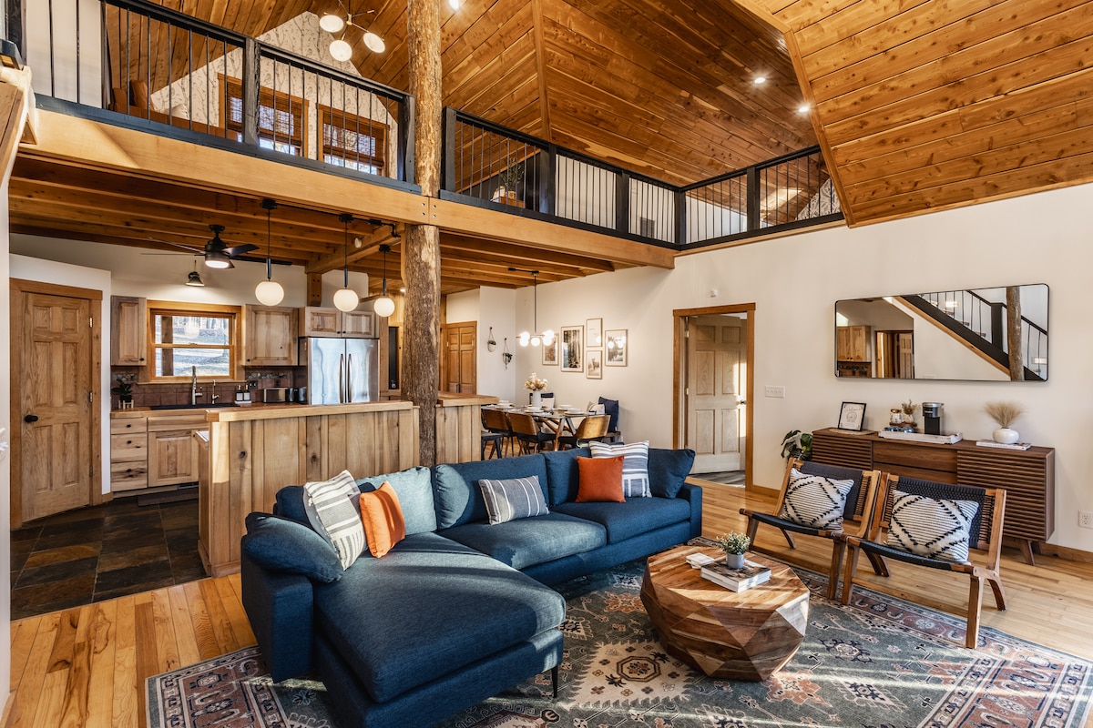 An open-concept living space with a high wooden ceiling features a comfortable blue sectional sofa, adorned with decorative pillows. A wooden coffee table is positioned on a patterned area rug, with a dining area visible in the background, showcasing a wooden table surrounded by chairs.