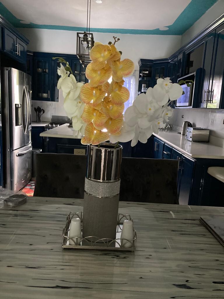 A dining table features a centerpiece of vibrant yellow and white orchids in an elegant vase. The background showcases a modern kitchen with dark blue cabinetry, stainless steel appliances, and a spacious countertop.