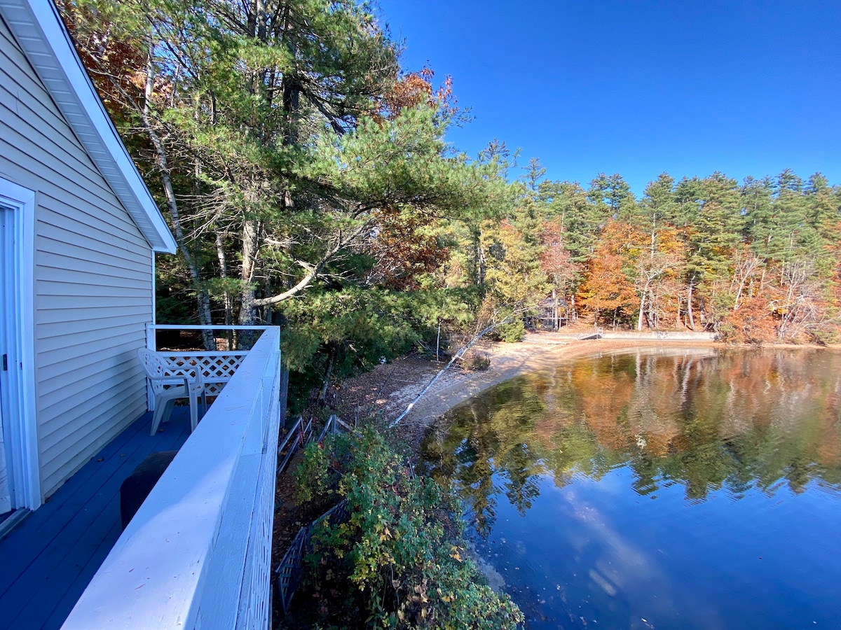 A serene view of a lakeside scene includes a wooden deck with a white chair overlooking calm water. Vibrant autumn foliage frames the scene, while a sandy beach with a gradual slope is visible, inviting relaxing moments by the water's edge.