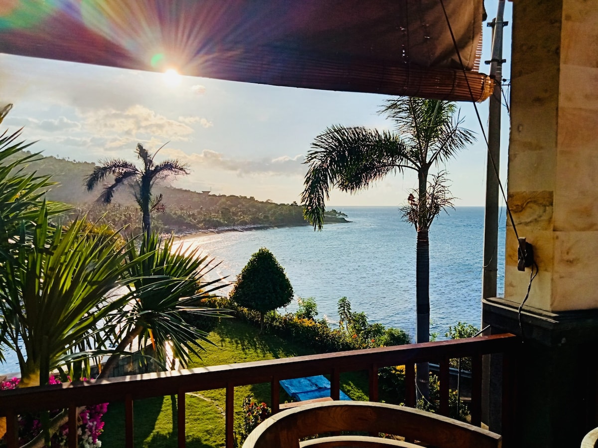 A sweeping view of the ocean and Lipah Beach is captured from a terrace. Lush greenery, including palm trees and flowering plants, frames the scene. Sunlight reflects off the water, creating a serene atmosphere, while distant coastal hills add depth to the landscape.