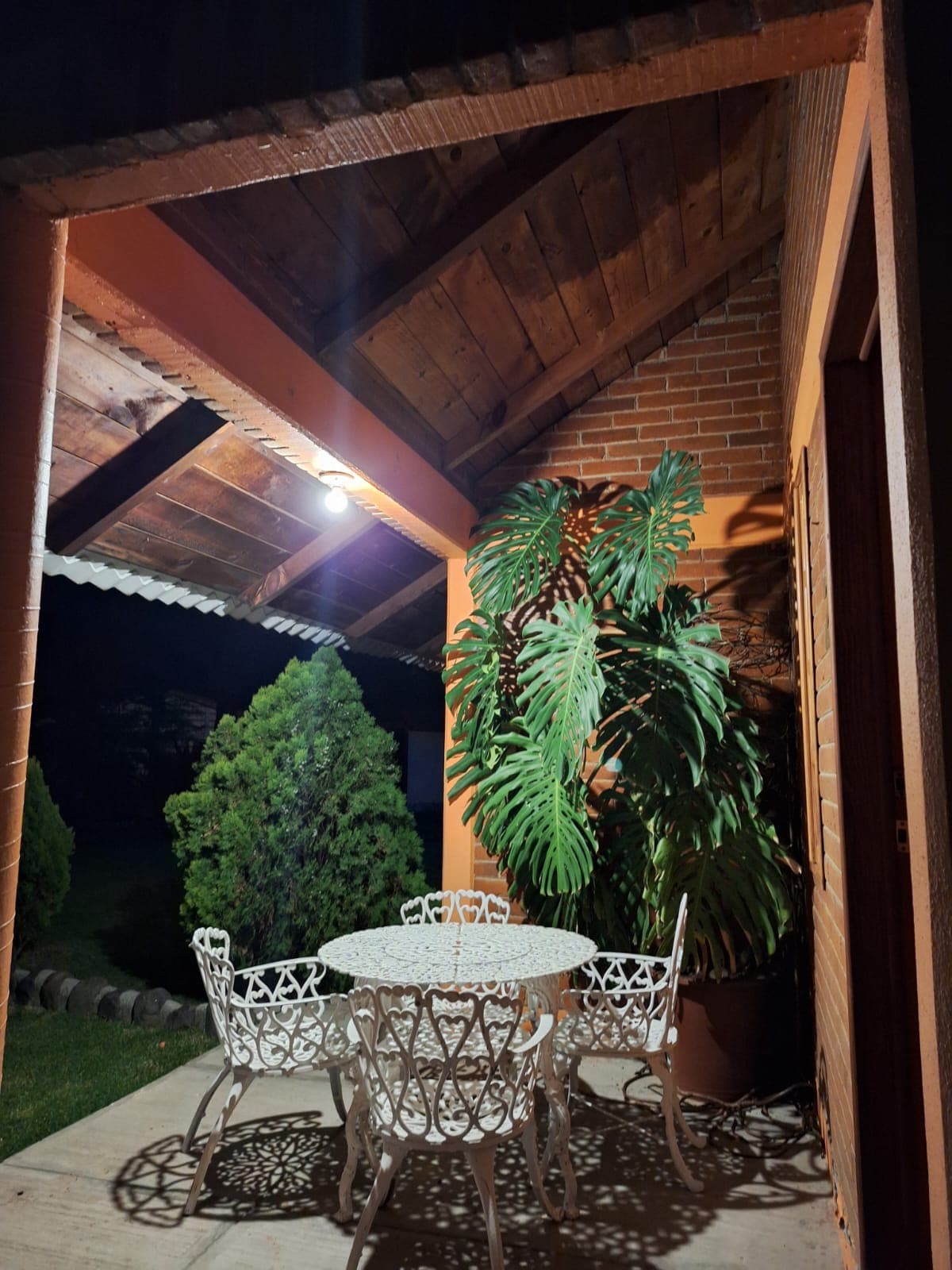 A charming outdoor patio area is illuminated by a soft light, featuring a round table surrounded by four intricate white chairs. Lush green plants add a touch of nature, while the sloped wooden ceiling enhances the cozy ambiance.