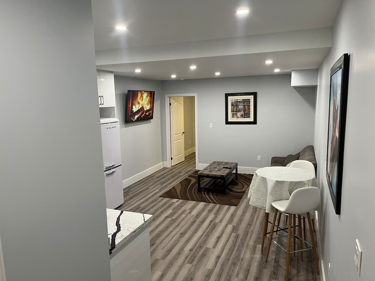 An open concept living space features a modern design with light grey walls and ample recessed lighting. A comfortable seating area is arranged beside a coffee table under a wall art piece. A small dining table next to a barstool is present, with a white kitchen in the background.