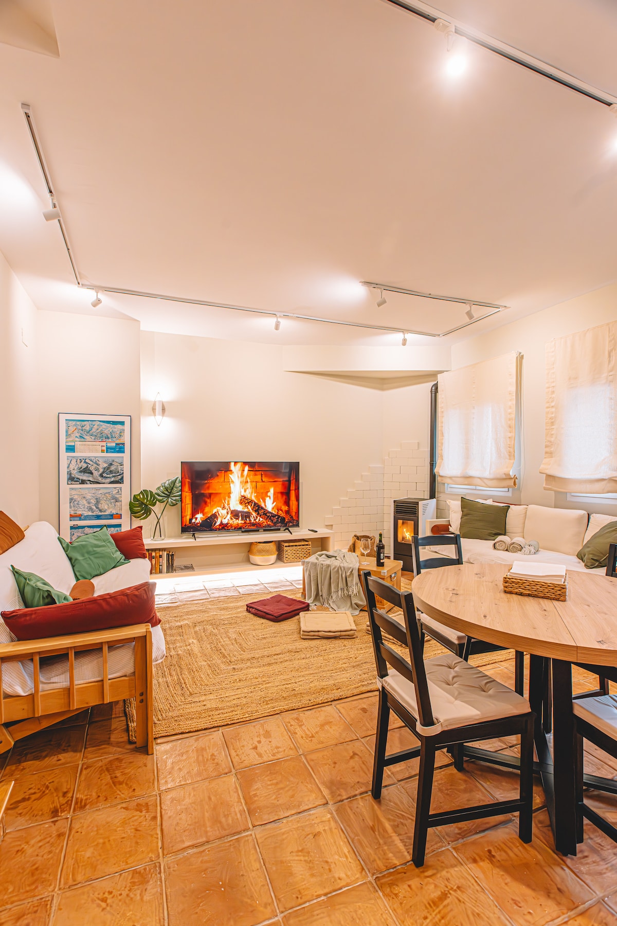 A welcoming living area features a cozy fireplace with a warm glow and a large screen TV displaying a fire. A round dining table is set with chairs, while a sofa is positioned nearby. Natural light filters through window coverings, enhancing the space's comfort.