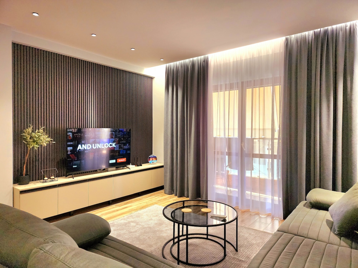 A modern living space features a sleek television mounted on a textured accent wall. Soft grey curtains frame large windows, allowing natural light to illuminate the room. A circular coffee table sits atop a light rug, accompanied by a plush sectional sofa, enhancing the cozy atmosphere.