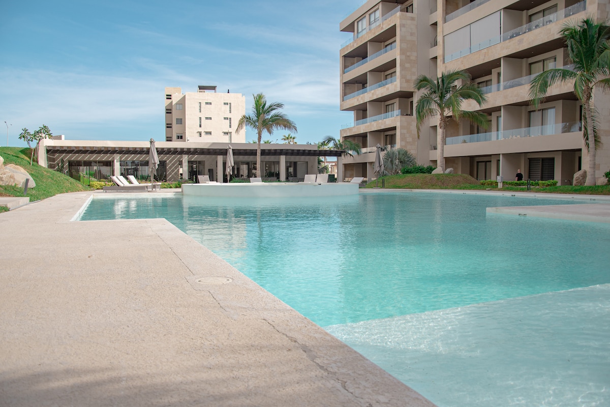 A serene pool area is surrounded by modern architecture and lush palm trees. The clear turquoise water reflects the sky, while a spacious lounge area with white sunbeds is visible in the background. The overall setting suggests a calm atmosphere for relaxation.
