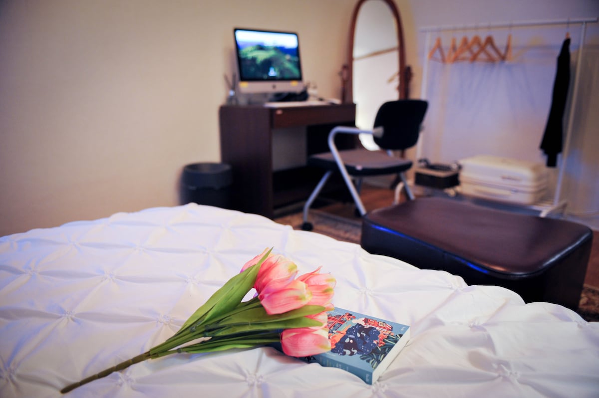 A comfortable sleeping area features a neatly made bed with a textured white coverlet. Nearby, a desk with a computer and an ergonomic chair is positioned against the wall. A bouquet of pink tulips and a book rest on the bed, adding a touch of color.