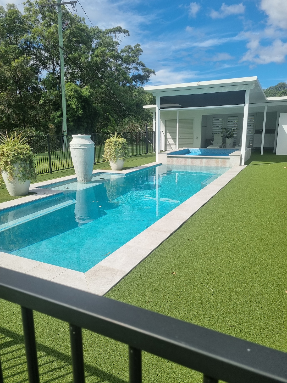 A sleek outdoor pool is surrounded by lush green lawn and modern landscaping. A fountain is positioned near the pool, and a shaded area with a heated spa is visible in the background. Clear blue skies provide a bright atmosphere.
