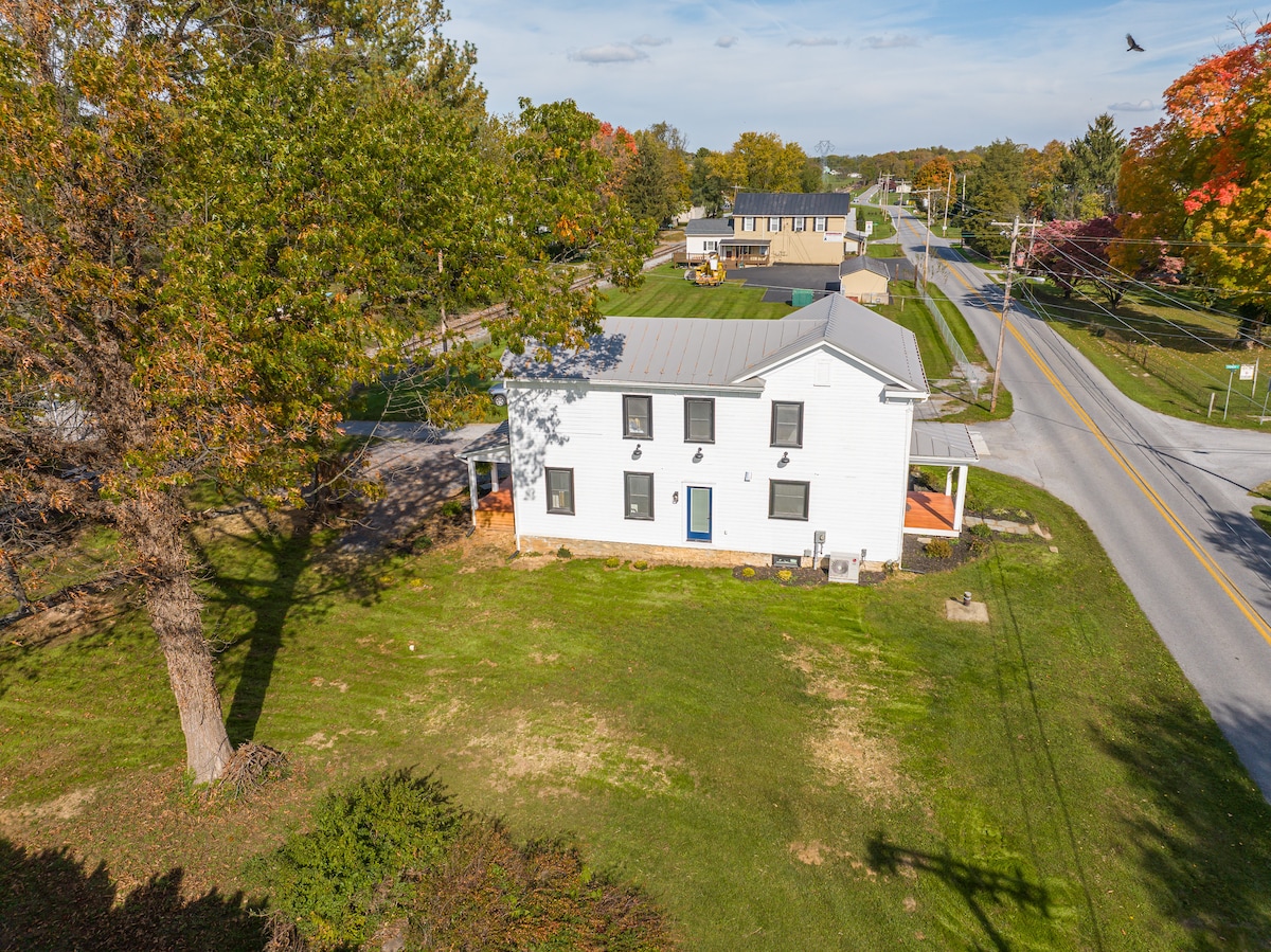 An aerial view of the historic home reveals its white exterior and well-maintained yard, surrounded by trees. The property sits at the intersection of two roads, showcasing the spacious front porch and the inviting entrance door. Nearby structures and roads are visible in the background.
