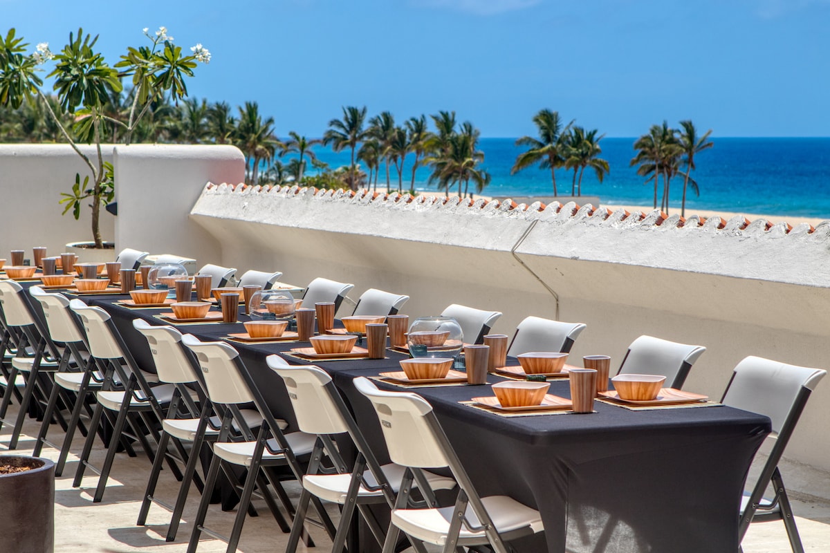 A long table set for a group meal is arranged on a terrace with a view of the ocean. Chairs are placed on either side of the table, which is adorned with bowls and drinkware. Palm trees line the coastline in the distant background under a clear blue sky.