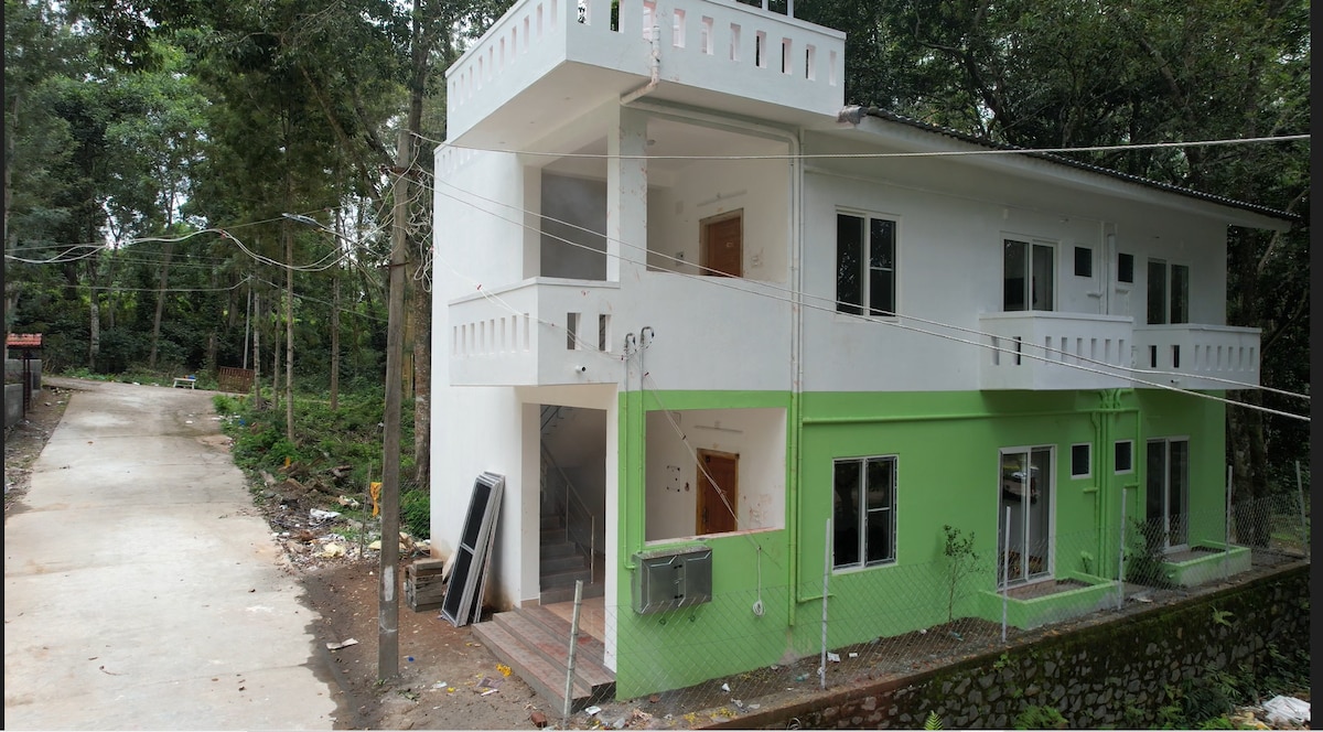 A modern two-story building is displayed, featuring a two-tone exterior with white and vibrant green walls. The structure is surrounded by lush greenery, and several windows are visible, allowing for natural light. The pathway in front leads to a wooded area.