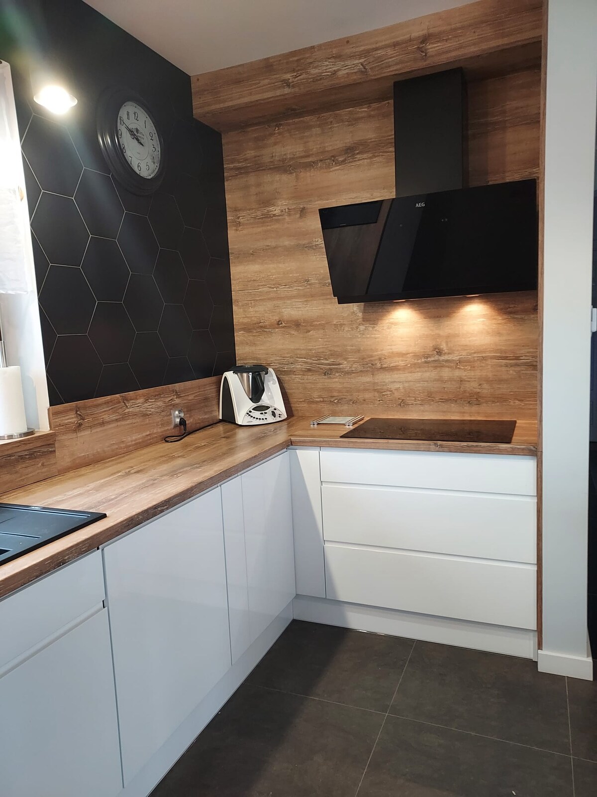 The kitchen features contemporary cabinetry with a mix of white and wooden finishes. A modern black extractor hood is mounted above a sleek cooking surface. Subtle lighting highlights the hexagonal black backsplash, while a clock adds a functional detail to the space.