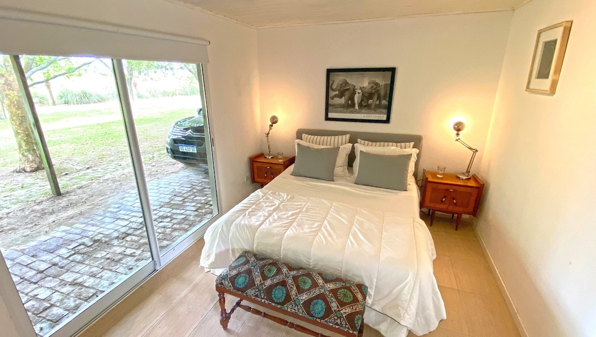 A serene bedroom is visible, featuring a spacious bed with gray and white bedding. Nightstands with lamps are positioned on either side, and a piece of artwork depicting elephants hangs above the bed. Large sliding doors open to a view of the exterior landscaping.