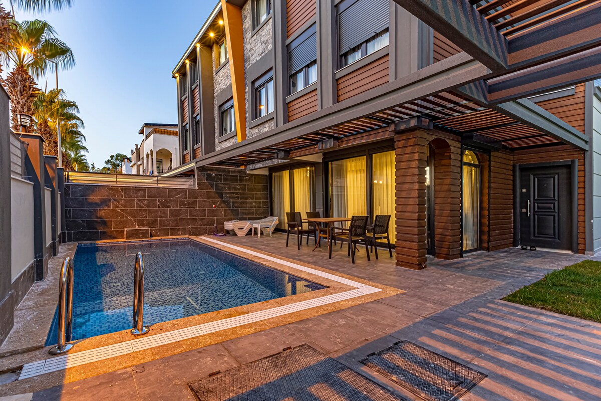 A private pool is featured in the spacious outdoor area, surrounded by a stone patio and lush greenery. Comfortable seating is arranged nearby under an overhead structure, enhancing the outdoor living experience. The villa's modern facade complements the tranquil setting.