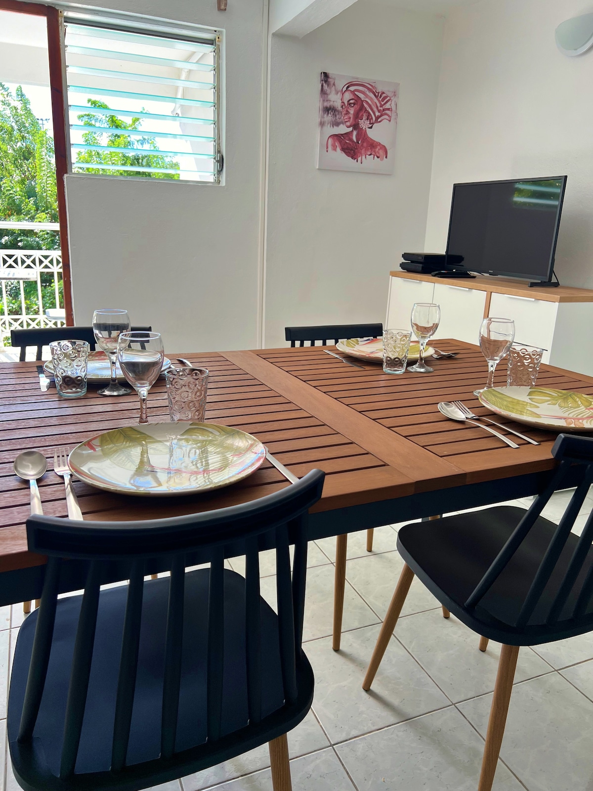 A dining area features a wooden table set with plates and glassware. Black chairs surround the table, creating a functional space for meals. A television is positioned nearby, while natural light enters through a window adorned with shutters.
