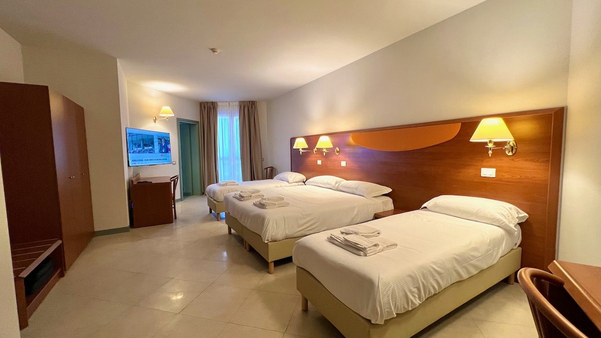 A spacious room is furnished with two beds and two single beds, accommodating various sleeping arrangements. Soft lighting is provided by wall-mounted lamps, while a flat-screen television is mounted on the wall. A large window offers natural light, complemented by light-colored curtains.