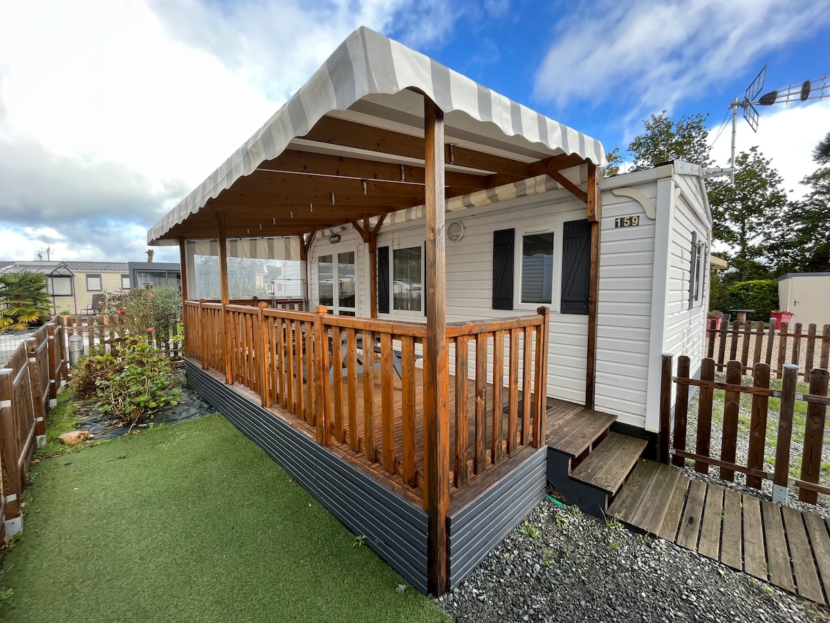 A mobile home is presented within a residential area, featuring a covered terrace with wooden railings. The exterior includes a well-kept lawn and a fenced yard, with cloudy skies visible above. The structure is oriented to create a welcoming outdoor space.