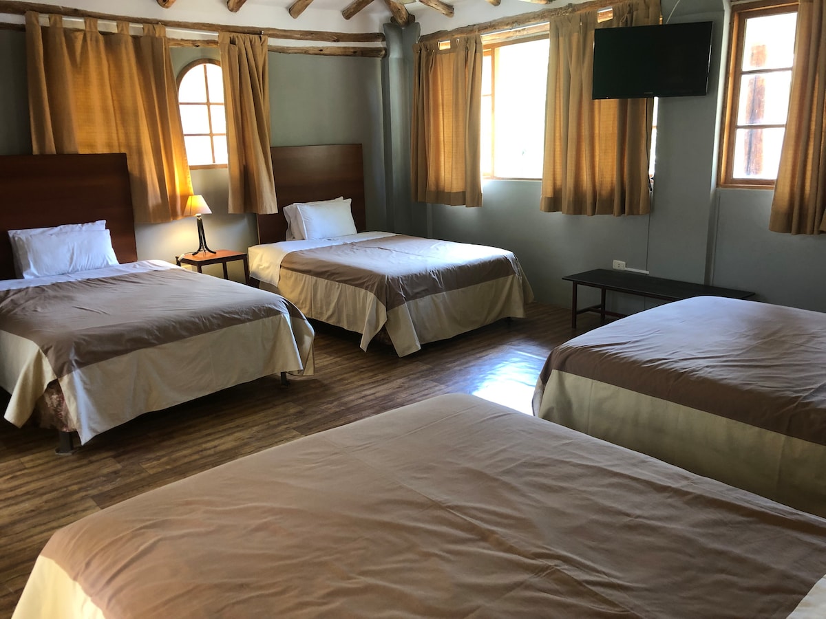 The sleeping area features multiple spacious beds arranged within a well-lit room. Light brown curtains frame large windows, enhancing natural light. A simple wooden table stands in the center, and a flat-screen TV is mounted on the wall.
