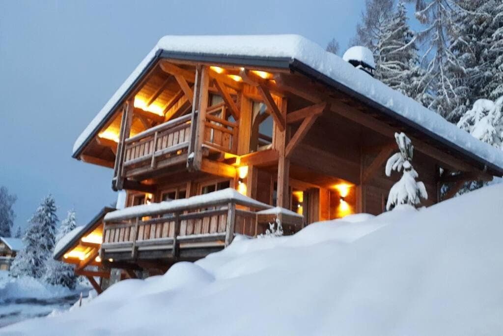 A wooden chalet nestled in a snowy landscape is illuminated by warm outdoor lights. Multiple balconies overlook the surrounding winter scenery, and fresh snow blankets the ground. The cozy structure exudes a rustic charm, enhanced by the gently sloping roof and wooden beams.