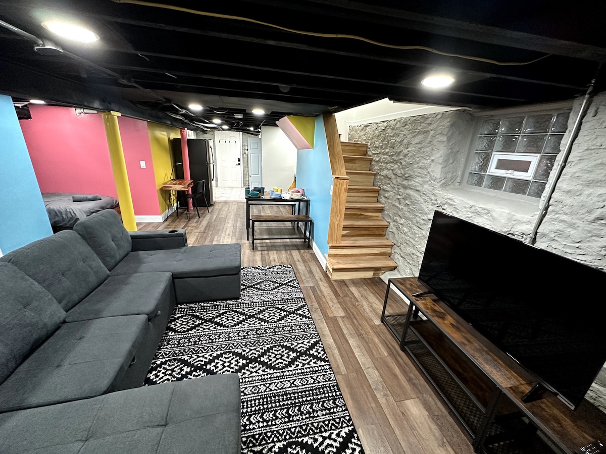 A spacious living area features a large, dark grey sectional sofa, a patterned rug, and a 55-inch television. The walls are painted in vibrant colors, and wooden stairs lead to an upper level. Natural light enters through glass-block windows, enhancing the overall brightness.